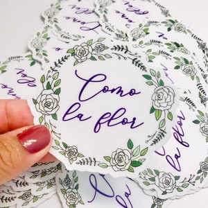 May include: A stack of white round stickers with a floral border and the text "Como la flor" in purple lettering.