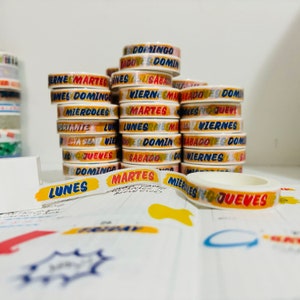 May include: A stack of colorful washi tapes with Spanish days of the week printed on them. The tapes are white with colorful lettering and illustrations. The tapes are arranged in a stack with the top tape showing the word "DOMINGO" in red and blue lettering.