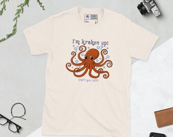 Puede incluir: Camiseta color crema con un diseño de pulpo de dibujos animados y el texto "I'm kraken up!" y "Can't you telly?" El pulpo es marrón con burbujas azules. La camiseta es de material suave.