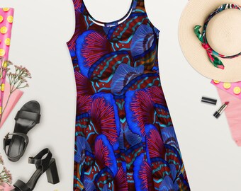 Op de afbeelding: Een mouwloze skaterjurk met een levendig, abstract patroon in tinten rood, blauw en blauwgroen. De jurk heeft een ronde hals en een uitlopende rok. De afbeelding bevat ook zwarte sandalen, een strohoed en make-up.