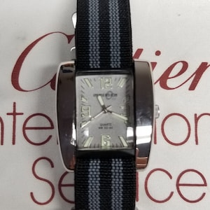 May include: A silver watch with a black and grey striped nylon strap. The watch face is rectangular and has a white background with black numbers and hands. The watch face also has the text "Invicta" and "Quartz WR 50 M" printed on it.