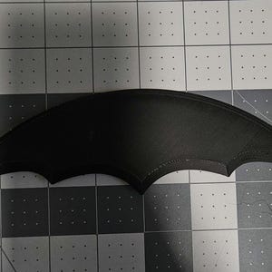 Puede incluir: Un aplique negro en forma de murciélago con una superficie texturizada. El aplique tiene una forma curva con bordes puntiagudos, que se asemejan a las alas de un murciélago. Se coloca sobre un fondo de cuadrícula gris y blanco.