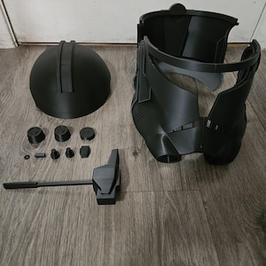Puede incluir: Un casco negro desmontado, probablemente para cosplay o disfraz, con varios componentes, incluyendo una cúpula, una máscara y piezas más pequeñas. Las piezas están dispuestas sobre una superficie de madera, sugiriendo un proyecto de bricolaje o un kit.