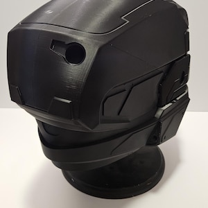 May include: A black, angular helmet with a matte finish, possibly for cosplay or a futuristic design. The helmet has a series of geometric panels and a circular opening on the front. It sits on a black, circular base.