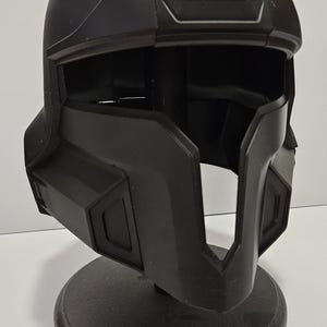 May include: A black, futuristic-looking helmet with a visor opening and geometric design. The helmet is displayed on a black stand, showcasing its angular features and overall shape. The helmet appears to be made of a matte material.