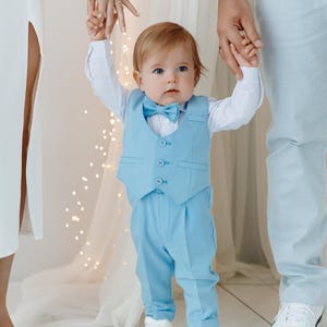 May include: A baby boy in a light blue suit with a bow tie is being held by his parents. He is wearing white shoes and has red hair.