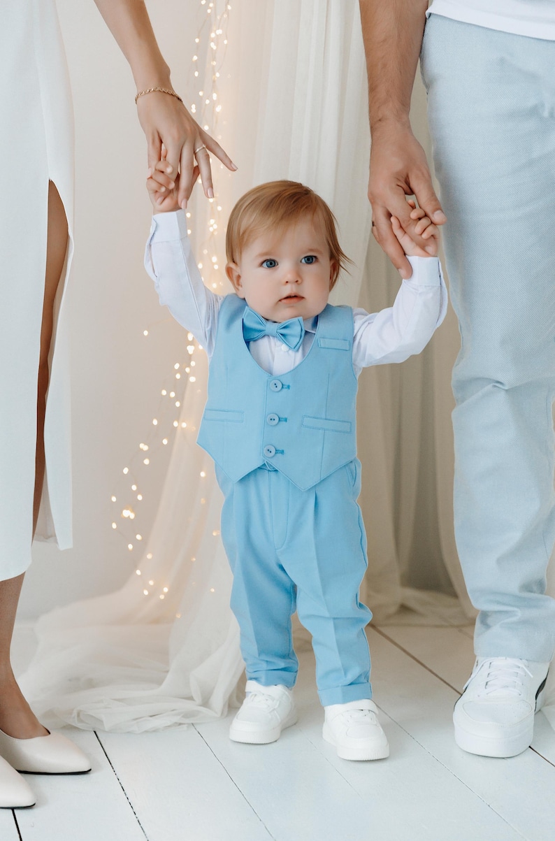 May include: A baby boy in a light blue suit with a bow tie is being held by his parents. He is wearing white shoes and has red hair.