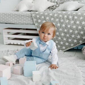 May include: A baby boy in a light blue suit and bow tie plays with wooden blocks on a white rug. The baby is sitting on the rug and is looking at the camera. There are other blocks scattered around the baby.
