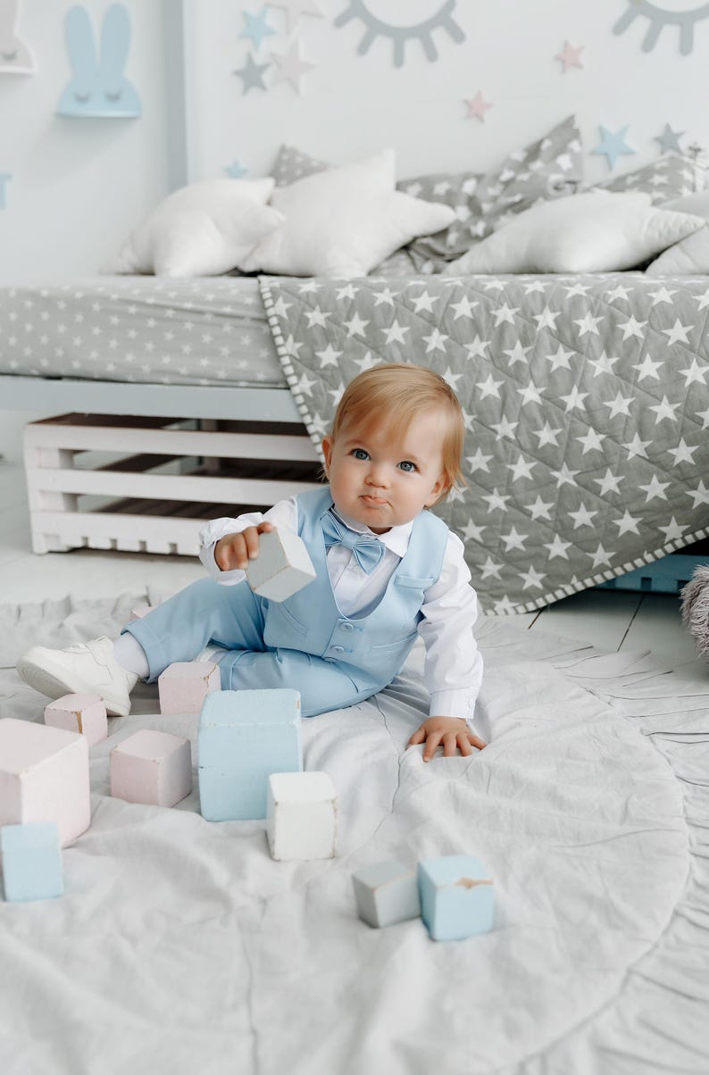 May include: A baby boy in a light blue suit and bow tie plays with wooden blocks on a white rug. The baby is sitting on the rug and is looking at the camera. There are other blocks scattered around the baby.