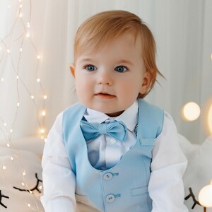 May include: A baby boy wearing a light blue vest with a matching bow tie. The vest has four buttons and the baby is sitting on a white surface with string lights in the background.