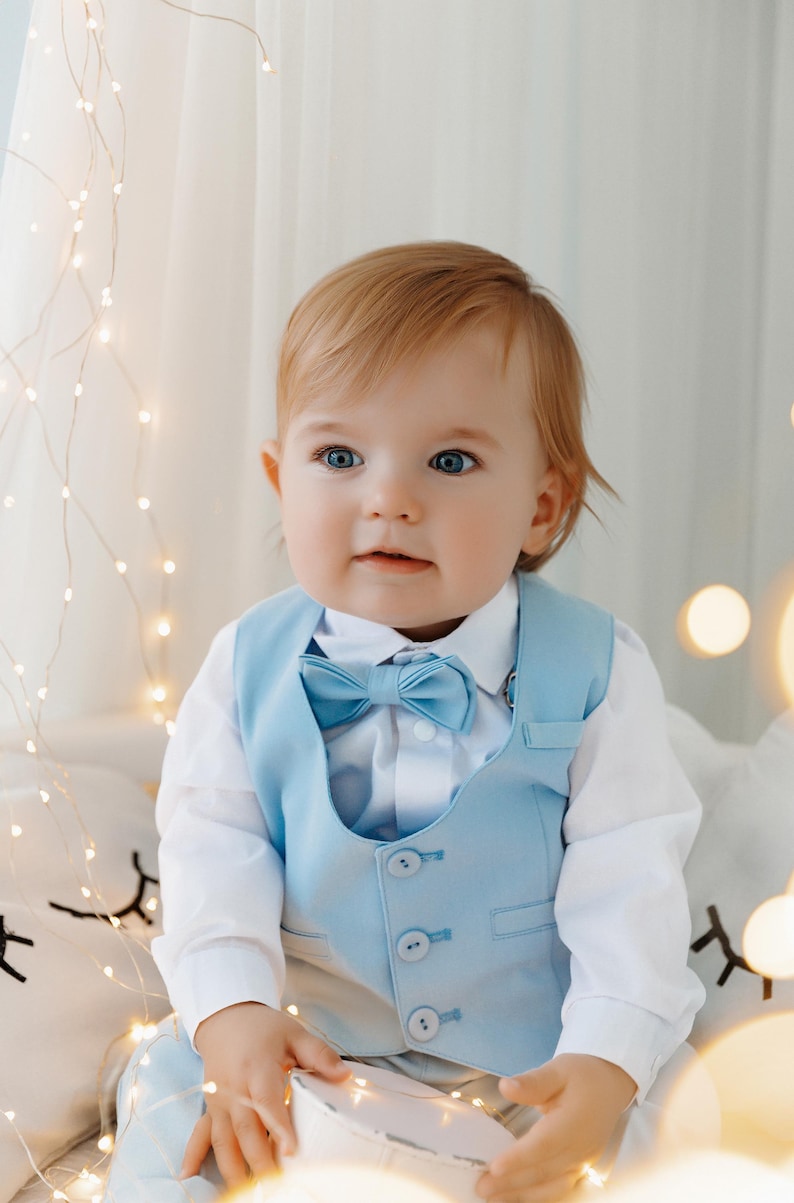 May include: A baby boy wearing a light blue vest with a matching bow tie. The vest has four buttons and the baby is sitting on a white surface with string lights in the background.