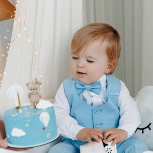 May include: A young child wearing a light blue suit with a bow tie looks at a blue and white birthday cake decorated with a teddy bear and balloons. The cake says "One" on it.