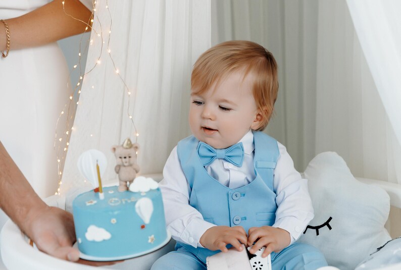 May include: A young child wearing a light blue suit with a bow tie looks at a blue and white birthday cake decorated with a teddy bear and balloons. The cake says "One" on it.