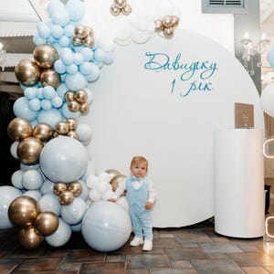 May include: A young boy in a light blue suit stands in front of a white backdrop decorated with blue and gold balloons. The backdrop has the text "Давидку 1 рік" written on it.