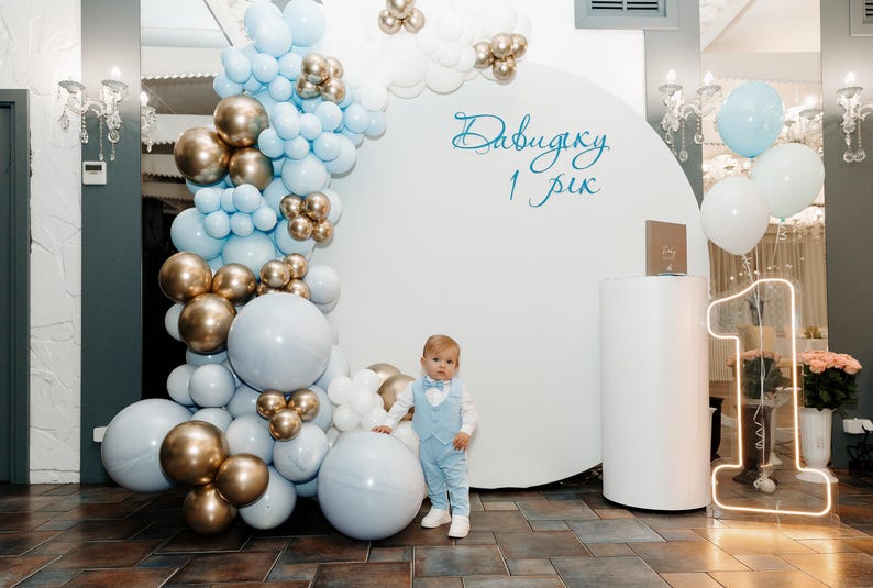 May include: A young boy in a light blue suit stands in front of a white backdrop decorated with blue and gold balloons. The backdrop has the text "Давидку 1 рік" written on it.