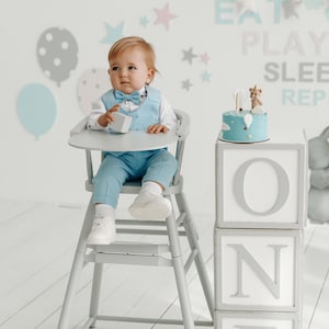May include: A baby boy in a light blue suit sits in a high chair in front of a cake decorated with a small bear figurine and the word "ONE" spelled out in large white letters on gray blocks.