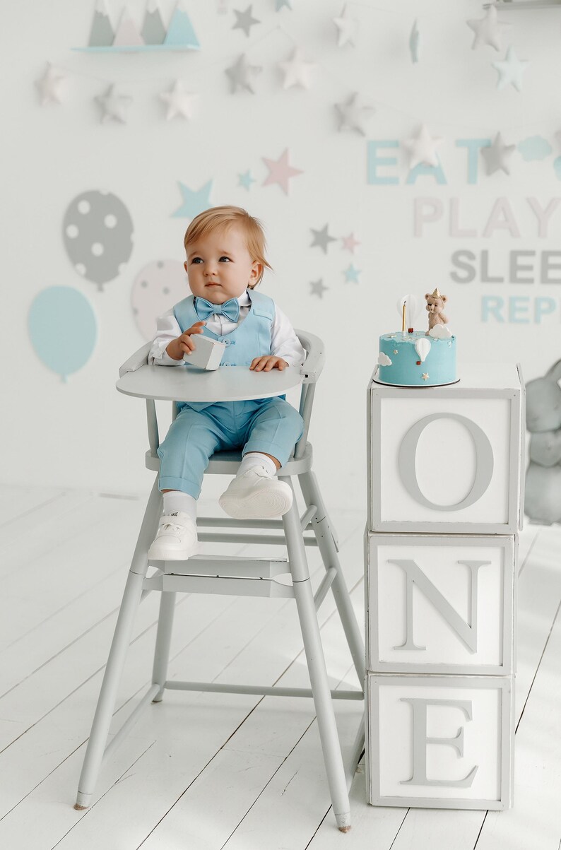 May include: A baby boy in a light blue suit sits in a high chair in front of a cake decorated with a small bear figurine and the word "ONE" spelled out in large white letters on gray blocks.