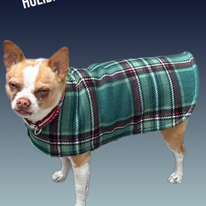 May include: A small dog wearing a green and red plaid fleece dog coat. The coat has a red and white trim. The dog is standing on a blue background. HOLIDAY 2023 is written in white text at the top of the image.