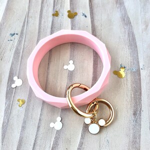 Puede incluir: Un brazalete rosa claro con un patr&oacute;n geom&eacute;trico, con un cierre y un llavero dorados. El llavero tiene un dije blanco con un contorno dorado. Confeti decorativo con forma de Mickey Mouse est&aacute; esparcido.