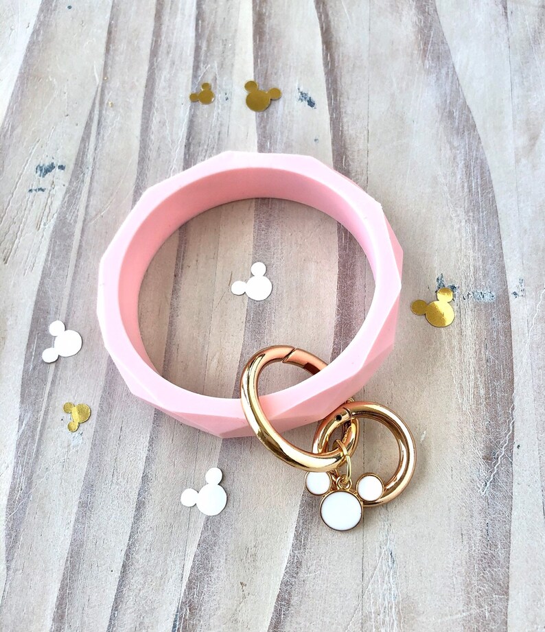 Puede incluir: Un brazalete rosa claro con un patr&oacute;n geom&eacute;trico, con un cierre y un llavero dorados. El llavero tiene un dije blanco con un contorno dorado. Confeti decorativo con forma de Mickey Mouse est&aacute; esparcido.
