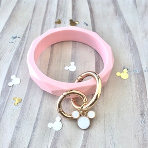 Puede incluir: Una pulsera brazalete rosa claro, facetada, con un cierre dorado y un charm con un dise&ntilde;o blanco de Mickey Mouse. La pulsera descansa sobre una superficie de madera clara, con confeti en forma de Mickey Mouse esparcido.