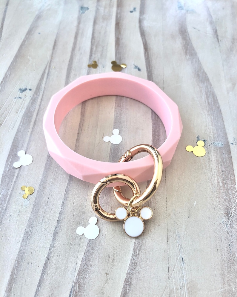 Puede incluir: Una pulsera brazalete rosa claro, facetada, con un cierre dorado y un charm con un dise&ntilde;o blanco de Mickey Mouse. La pulsera descansa sobre una superficie de madera clara, con confeti en forma de Mickey Mouse esparcido.