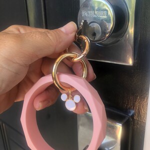 Puede incluir: Un llavero de pulsera de silicona rosa con un cierre dorado y un dije con un dise&ntilde;o de orejas de rat&oacute;n blanco y rosa. El llavero est&aacute; sujeto a una llave en una cerradura plateada.