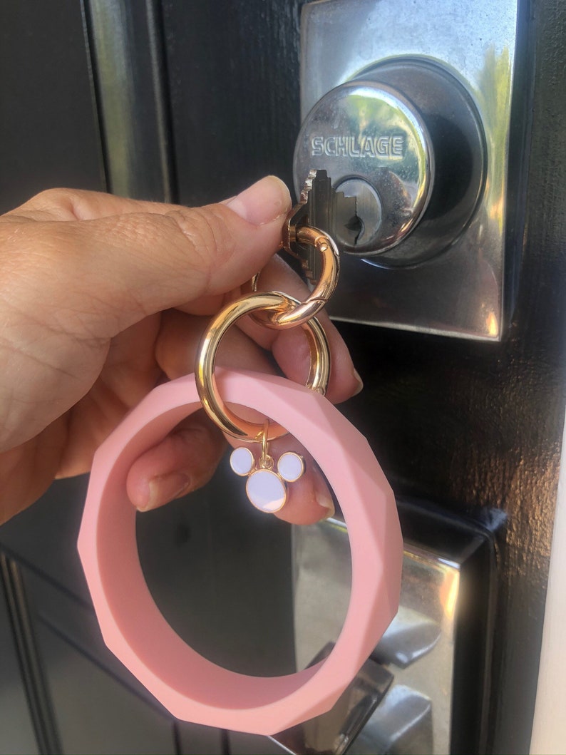 Puede incluir: Un llavero de pulsera de silicona rosa con un cierre dorado y un dije con un dise&ntilde;o de orejas de rat&oacute;n blanco y rosa. El llavero est&aacute; sujeto a una llave en una cerradura plateada.