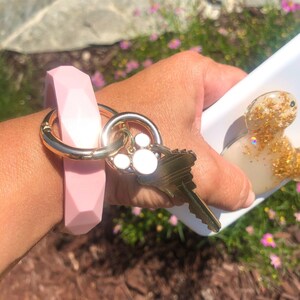 Puede incluir: Un llavero de pulsera de silicona rosa claro con un anillo de metal dorado y una llave. El llavero tiene un dise&ntilde;o geom&eacute;trico y est&aacute; unido a un llavero dorado con un dije de huella de pata.