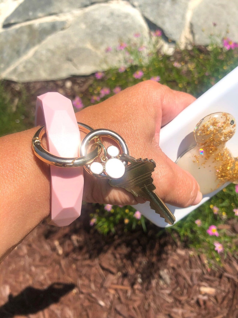 Puede incluir: Un llavero de pulsera de silicona rosa claro con un anillo de metal dorado y una llave. El llavero tiene un dise&ntilde;o geom&eacute;trico y est&aacute; unido a un llavero dorado con un dije de huella de pata.