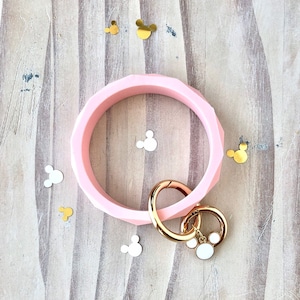 Puede incluir: Un brazalete de silicona rosa claro con un dise&ntilde;o geom&eacute;trico. Tiene un cierre dorado y un charm con una cabeza de Mickey Mouse blanca. Confeti de Mickey Mouse dorado y blanco est&aacute; esparcido.