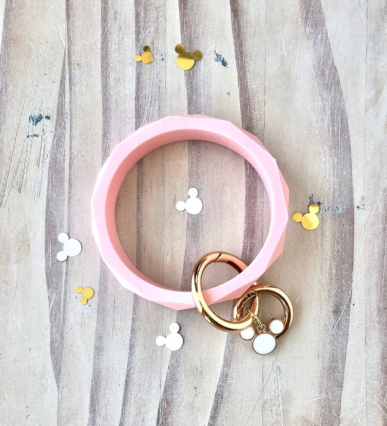 Puede incluir: Un brazalete de silicona rosa claro con un dise&ntilde;o geom&eacute;trico. Tiene un cierre dorado y un charm con una cabeza de Mickey Mouse blanca. Confeti de Mickey Mouse dorado y blanco est&aacute; esparcido.