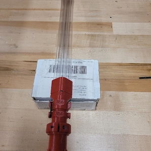 May include: A red 3D-printed object with a clear tube, possibly a tool or part. The object has a cylindrical design with the numbers "356" printed on it. It is resting on a cardboard box on a wooden surface.