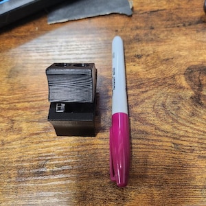 May include: A black plastic electrical terminal block with three screw terminals. The terminal block is sitting on a wooden surface next to a pink marker.