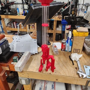 May include: A red and clear plastic reloading tool with a funnel on top. The tool is sitting on a wooden workbench with other tools and supplies.