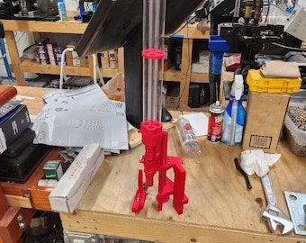 May include: A red and clear plastic reloading tool with a funnel on top. The tool is sitting on a wooden workbench with other tools and supplies.