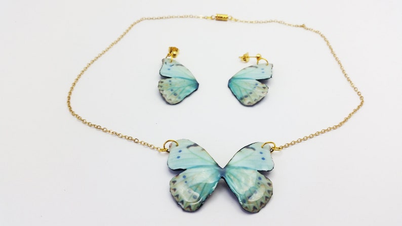 May include: A delicate gold chain necklace and matching earrings feature a light blue butterfly design. The butterfly wings have a subtle white and gray pattern.