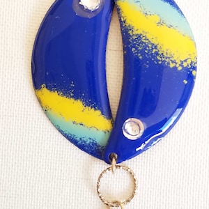 Peut inclure: Un pendentif en émail bleu et jaune avec deux strass transparents. Le pendentif est attaché à une chaîne dorée avec une petite goutte d'émail bleu.