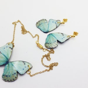 May include: A set of butterfly jewelry featuring a necklace and earrings. The butterfly wings are made of blue and green iridescent material and are attached to gold chains and earring posts.
