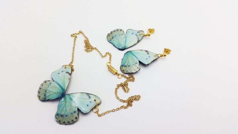 May include: A set of butterfly jewelry featuring a necklace and earrings. The butterfly wings are made of blue and green iridescent material and are attached to gold chains and earring posts.