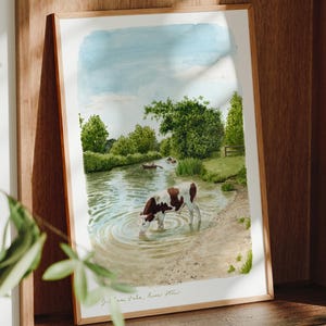 Puede incluir: Una impresión en acuarela enmarcada representa una serena escena de río con una vaca vadeando el agua. La obra de arte presenta un cielo azul claro, árboles verdes y una orilla arenosa. La impresión se exhibe en un estante de madera.