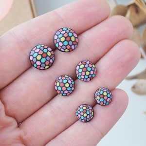 Könnte beinhalten: Sechs runde Ohrstecker mit einem bunten Mosaikmuster. Die Ohrringe haben eine schwarze Basis und ein Design aus kleinen, unregelmäßigen Formen in Rosa, Gelb, Blau und Grün. Die Ohrringe werden auf einer Hand gezeigt.
