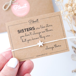 May include: A white string bracelet with a star charm is held in front of a card that reads "SISTERS are like stars, you don't always see them, but you know they are... always there." A small brown gift box is in the background.