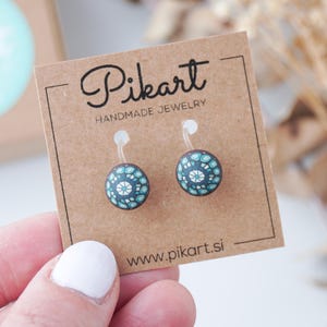 Può includere: Orecchini a bottone rotondi con un motivo floreale blu scuro e bianco. Gli orecchini sono esposti su una scheda marrone con il testo "Pikart Handmade Jewelry" e l'indirizzo del sito web www.pikart.si.