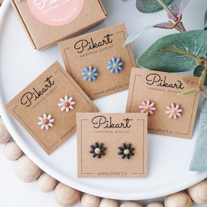 Peut inclure: Diverses boucles d'oreilles clous en forme de fleurs faites à la main, de couleurs blanche, bleu clair, rose et noire, présentées sur une assiette blanche. Chaque paire est sur une petite carte avec le logo "Pikart Handmade Jewelry".