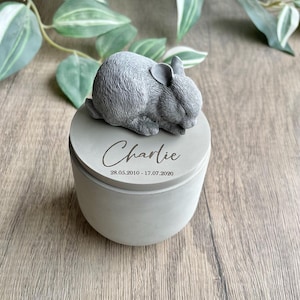 Peut inclure: Une boîte à souvenirs en béton gris avec une figurine de lapin endormi sur le couvercle. Le couvercle est gravé du nom "Charlie" et des dates "28.05.2010 - 17.07.2020".