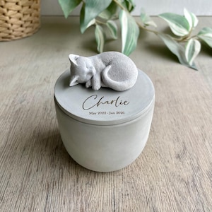 May include: A light gray concrete cylindrical urn with a removable lid featuring a sculpted sleeping cat. The lid is engraved with the name Charlie and the dates May 2022 - Jan 2026 in a dark script and sans-serif font combination.