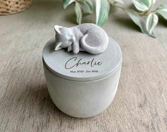 May include: A light gray concrete cylindrical urn with a removable lid featuring a sculpted sleeping cat. The lid is engraved with the name Charlie and the dates May 2022 - Jan 2026 in a dark script and sans-serif font combination.
