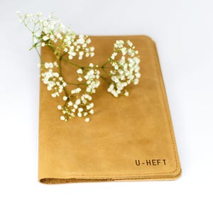 Könnte beinhalten: Eine U-Hefthülle aus Leder in Amaretti mit dem geprägten Text "U-HEFT" auf dem vorderen Umschlag. Ein Zweig weißer Gipskrautblüten liegt auf dem Umschlag.