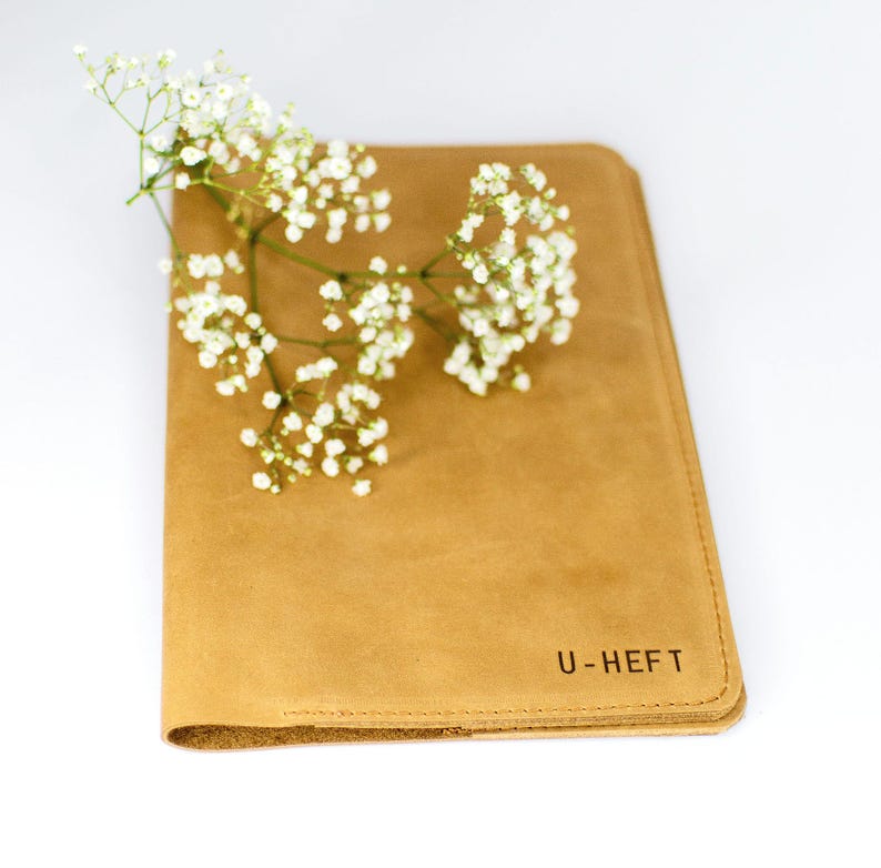 Könnte beinhalten: Eine U-Hefthülle aus Leder in Amaretti mit dem geprägten Text "U-HEFT" auf dem vorderen Umschlag. Ein Zweig weißer Gipskrautblüten liegt auf dem Umschlag.